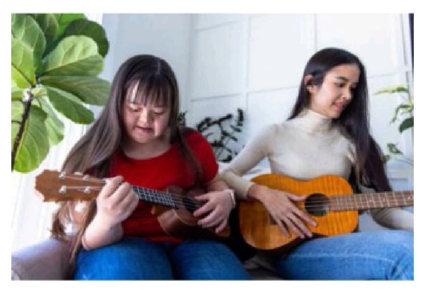 Two young women sitting on a couch playing guitars, surrounded by indoor plants in a bright room.